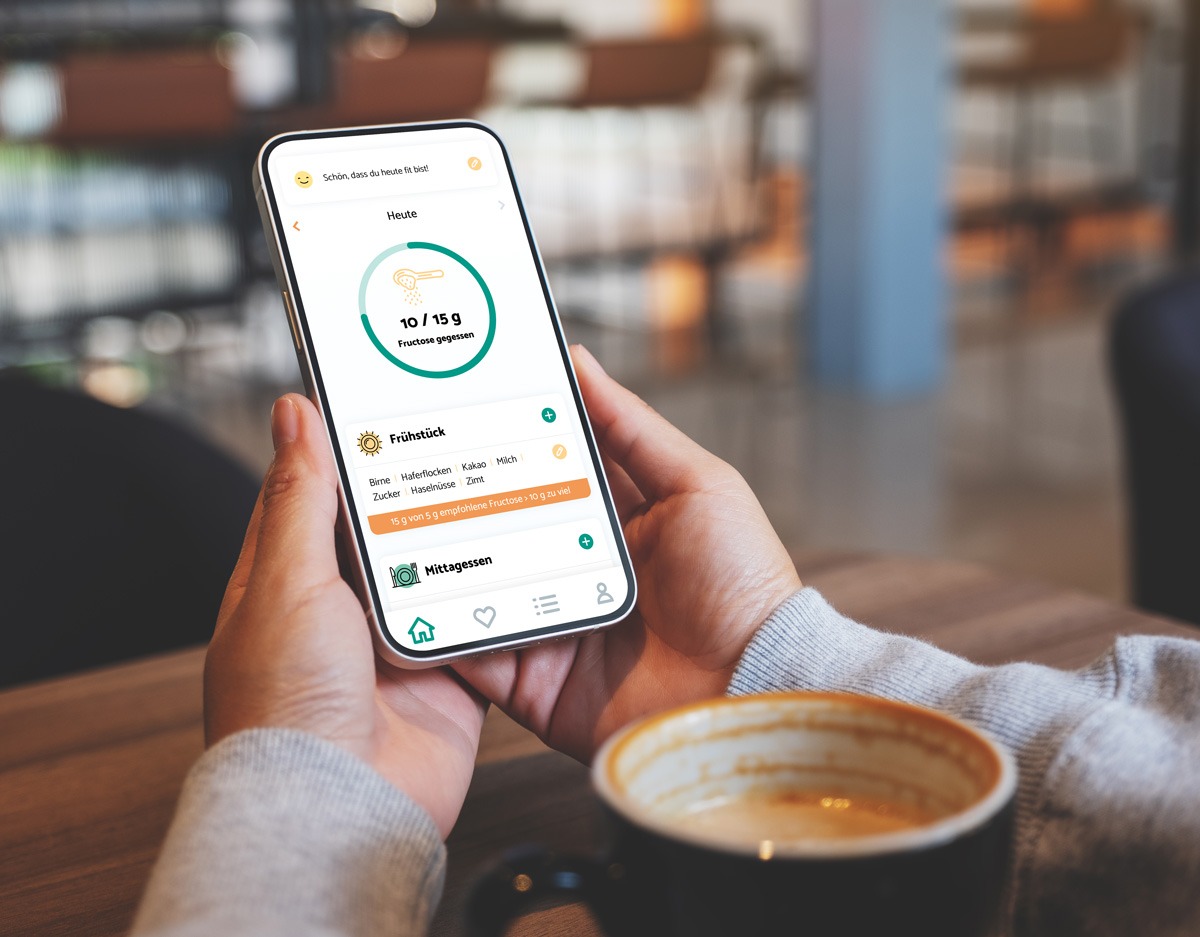 Frau hält ein Smartphone mit beiden Händen. Auf dem Bildschirm ist die Startseite der App zu sehen. Im Vordergrund sieht man eine Tasse mit Kaffee.